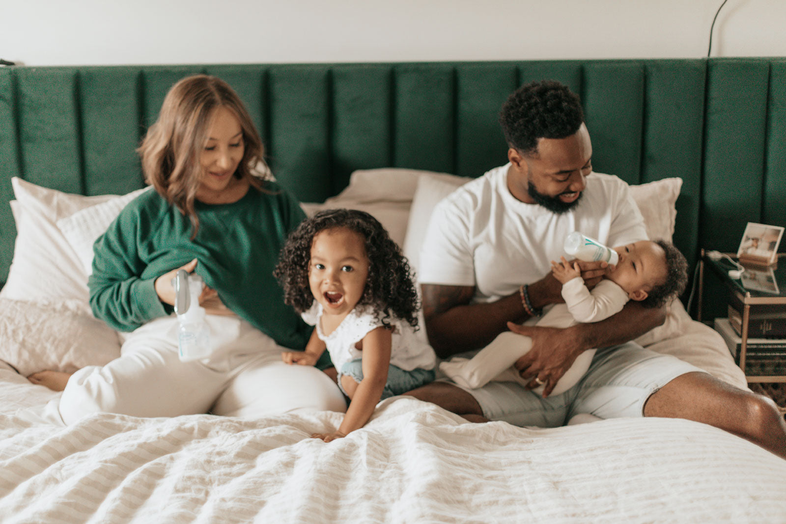 Family sitting in bed together with father bottle feeding and mother pumping