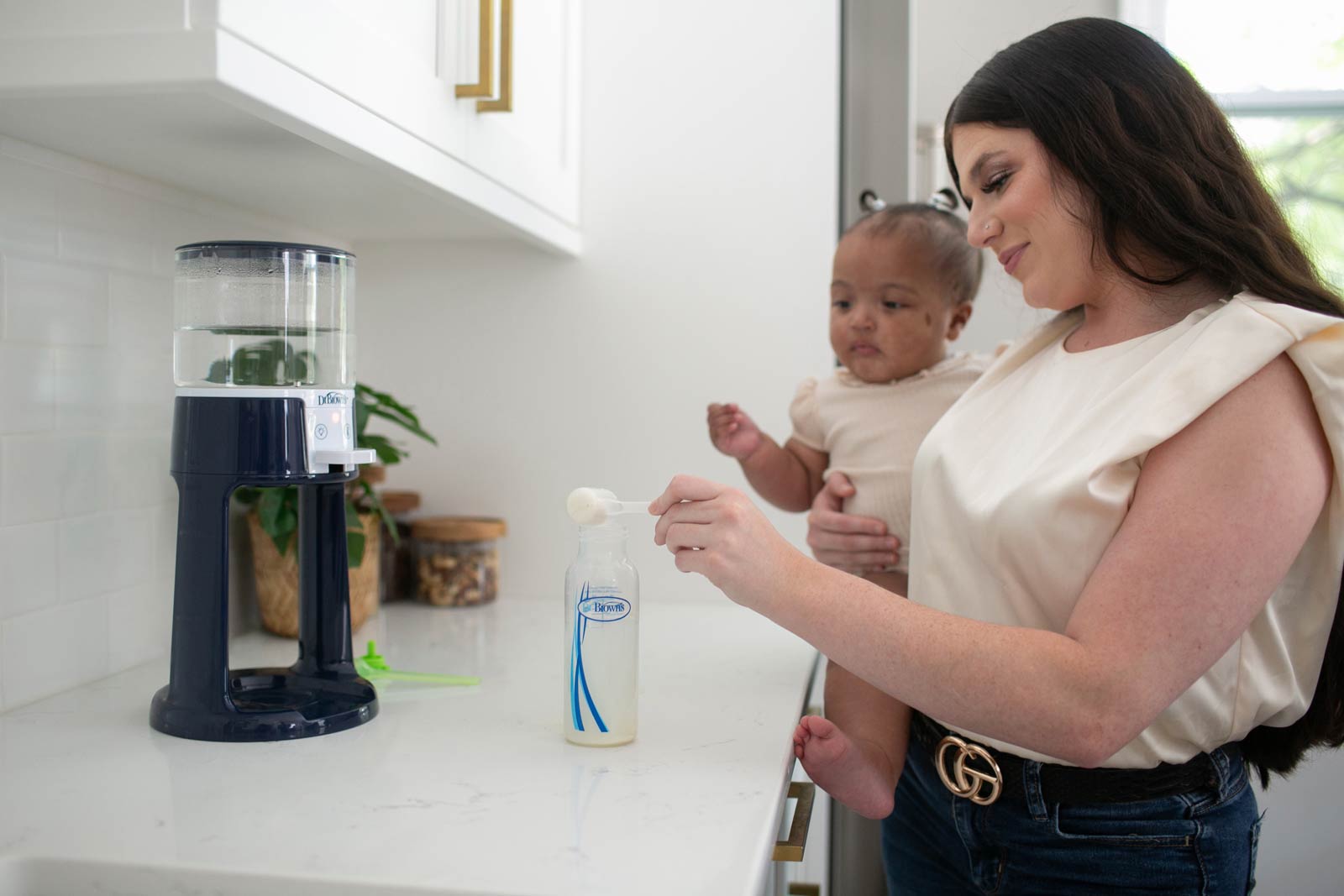 Mother holding baby and putting formula into bottle on a kitchen counter