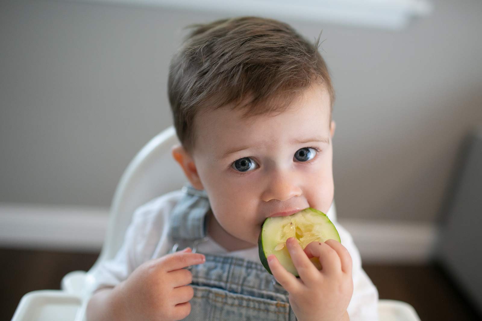 Toddler in a high chair biting into a cucumber