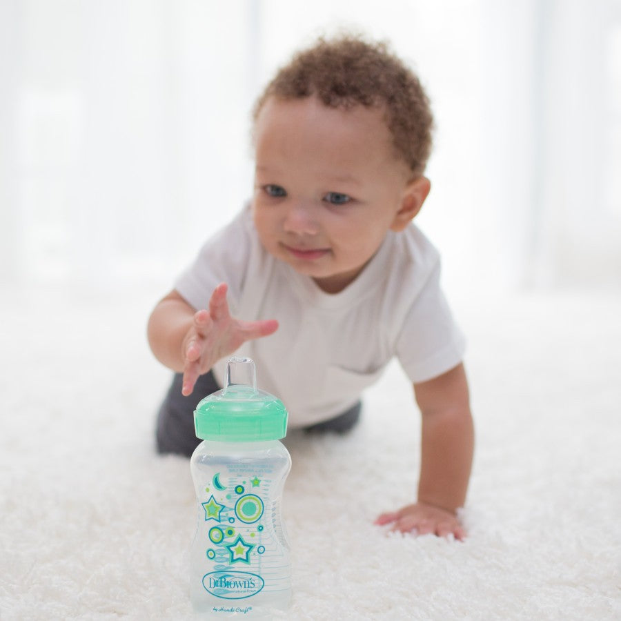 A baby in a white shirt is crawling on a white carpet, reaching for a green and clear baby bottle decorated with star and circle patterns. The room is softly lit.