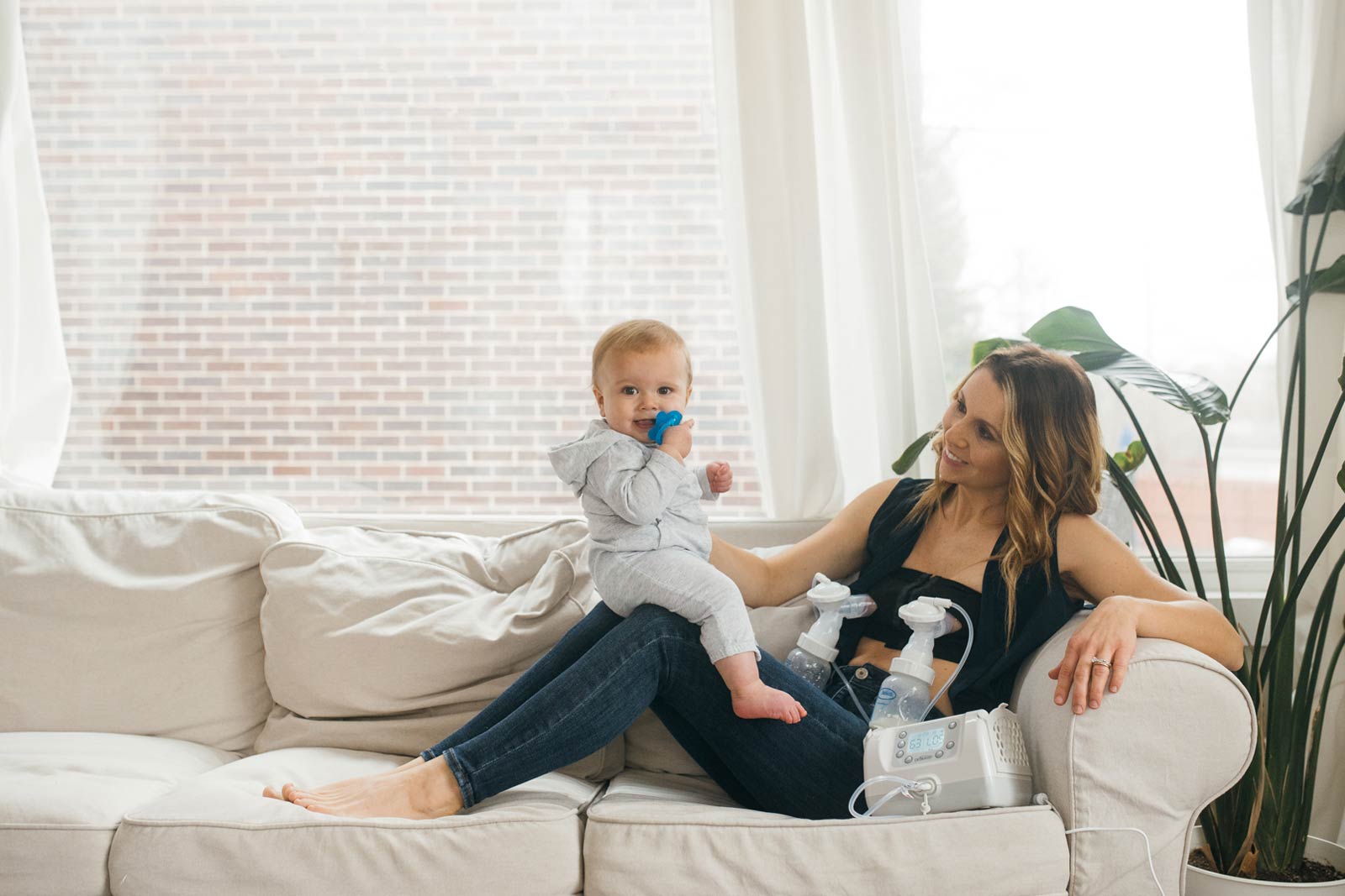 A woman is sitting on a sofa with a baby on her lap. The baby is holding a blue toy. Next to her is a breast pump and accessories. The room has large windows with light curtains and a view of a brick wall outside.
