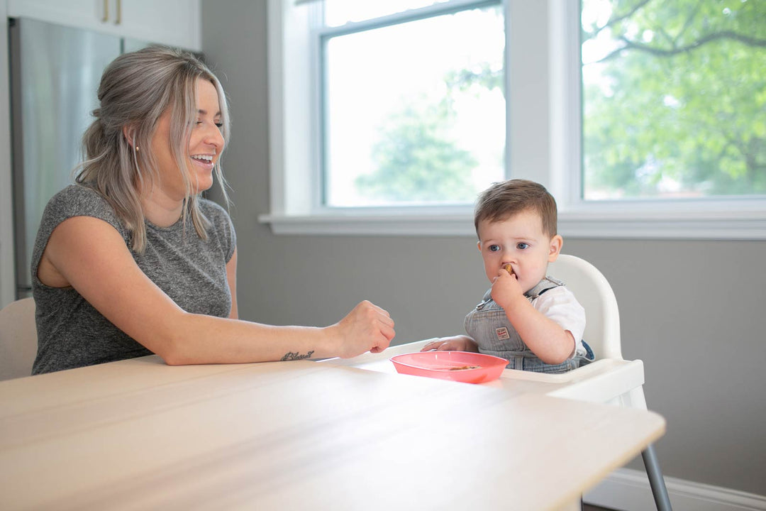 Mother and toddler sitting at a table, with the toddler in a high chair eating solid foods