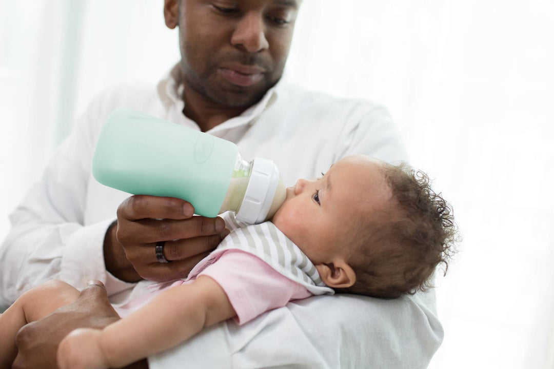 A man in a white shirt holds a baby wearing a pink and white striped outfit while feeding her with a mint green bottle.