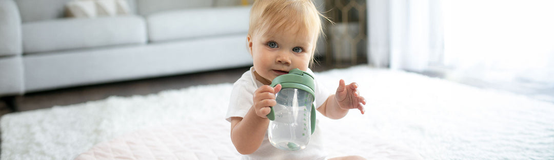 Baby drinking from a green Dr. Brown's Baby's First Straw Cup while seated on a white rug.