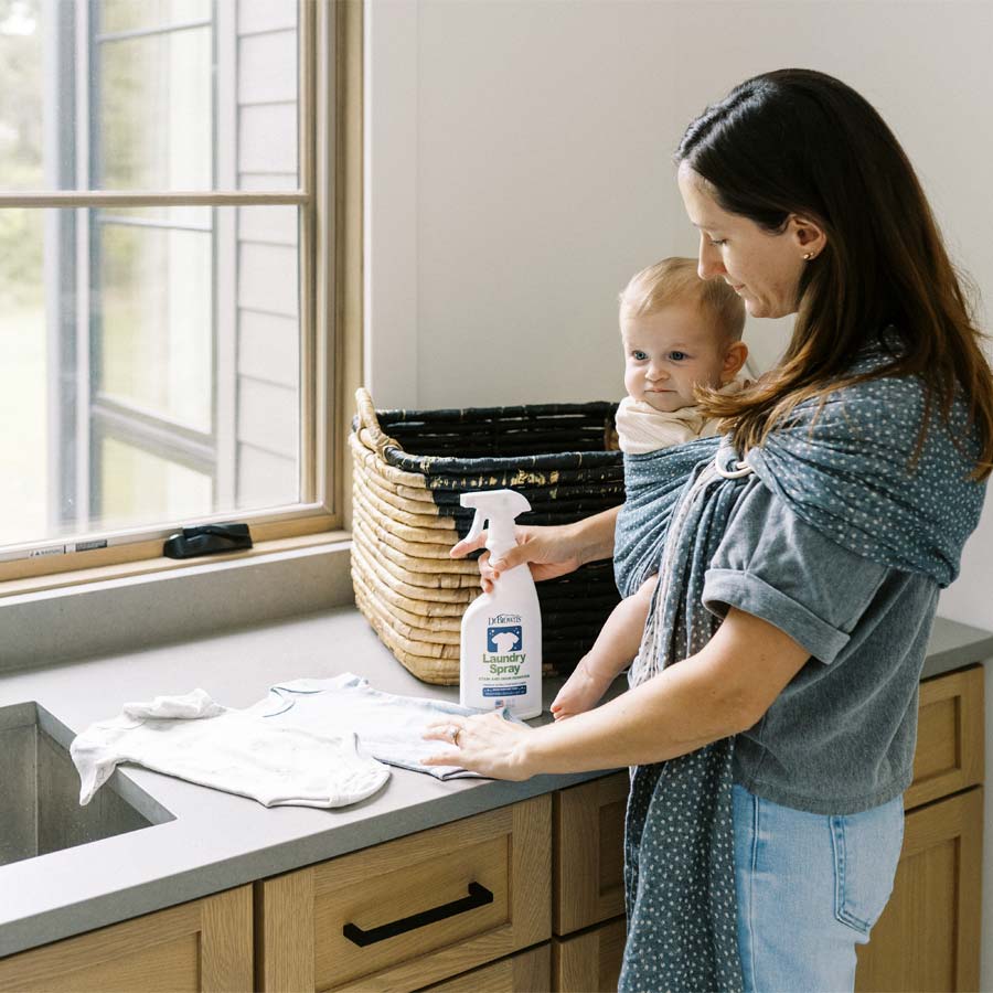 A woman holding a baby in a sling stands in a kitchen near a window. She sprays a cloth with a cleaning solution. A woven basket sits on the counter next to them. The setting is bright and tidy, creating a calm atmosphere.