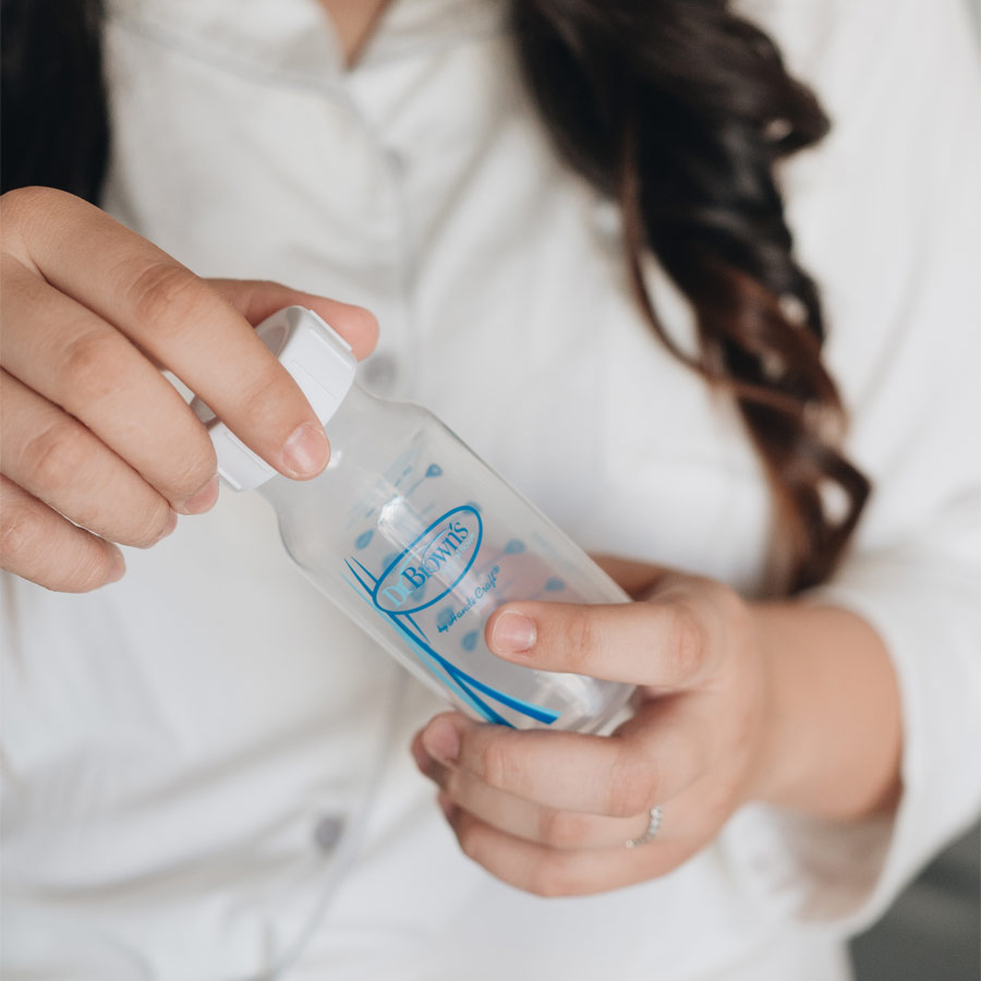 A person wearing a white shirt holds a small, clear baby bottle with a white cap and blue design. Their hands are in focus, showing neatly trimmed nails, and part of their long dark hair is visible.