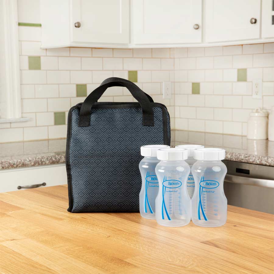 A black insulated bag with a handle sits on a wooden kitchen counter, alongside four empty, clear plastic baby bottles with white caps. The kitchen features a tiled backsplash and stainless steel appliances.