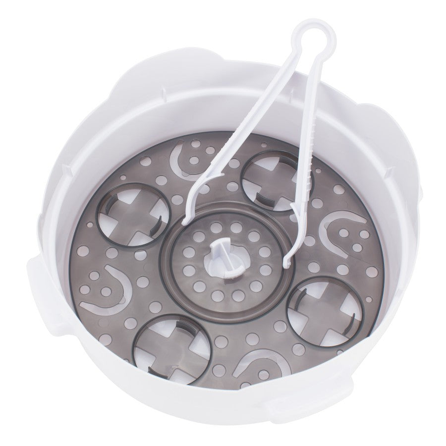 A white plastic steamer basket with a gray circular base featuring various cut-out shapes, including smiley faces and crosses. It includes a pair of tongs resting inside.