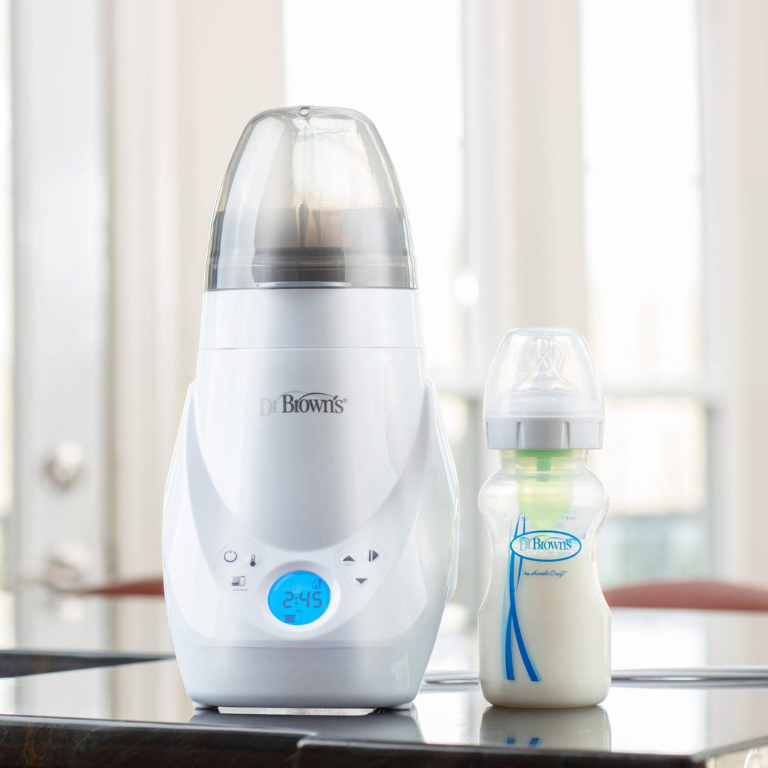 A Dr. Brown's baby bottle warmer with digital display sits on a table next to a filled baby bottle. The warmer shows 2:45 on the screen, and the background features large, bright windows.