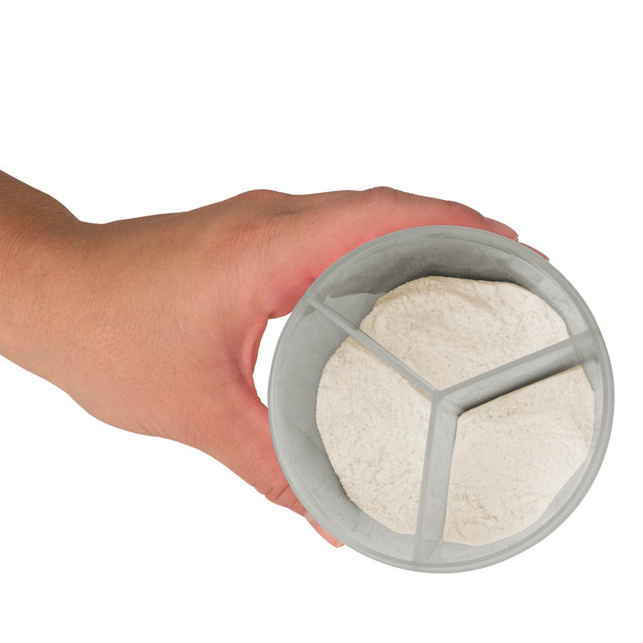 A hand holding a round gray formula dispenser divided into three compartments, filled with a white powder.