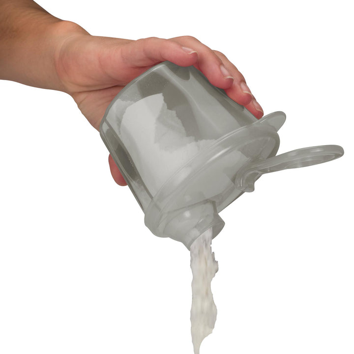 A hand holding a translucent gray formula dispenser with a lid and spout, pouring a white creamy substance into a stream.