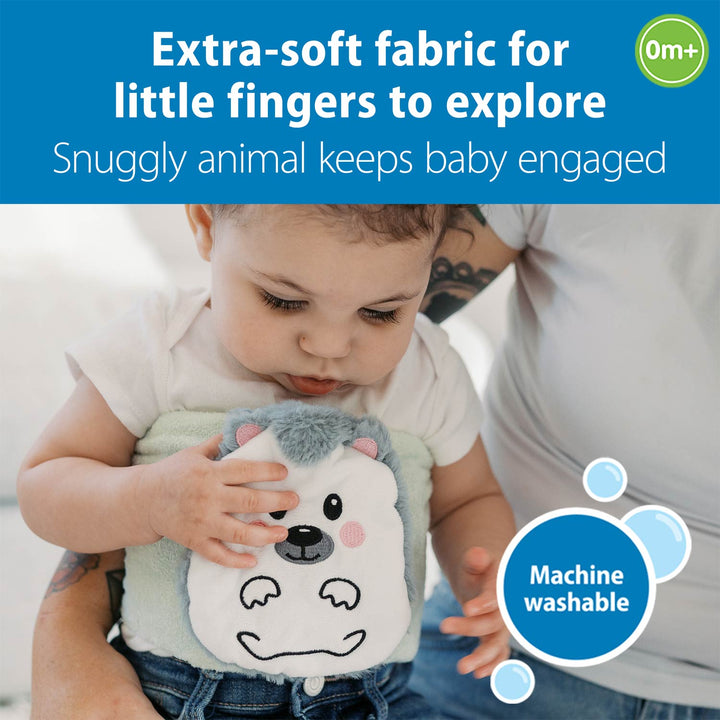 A baby in a white onesie is being held by an adult while touching a plush animal belly band. The belly band is light green with a white bear face and gray ears.