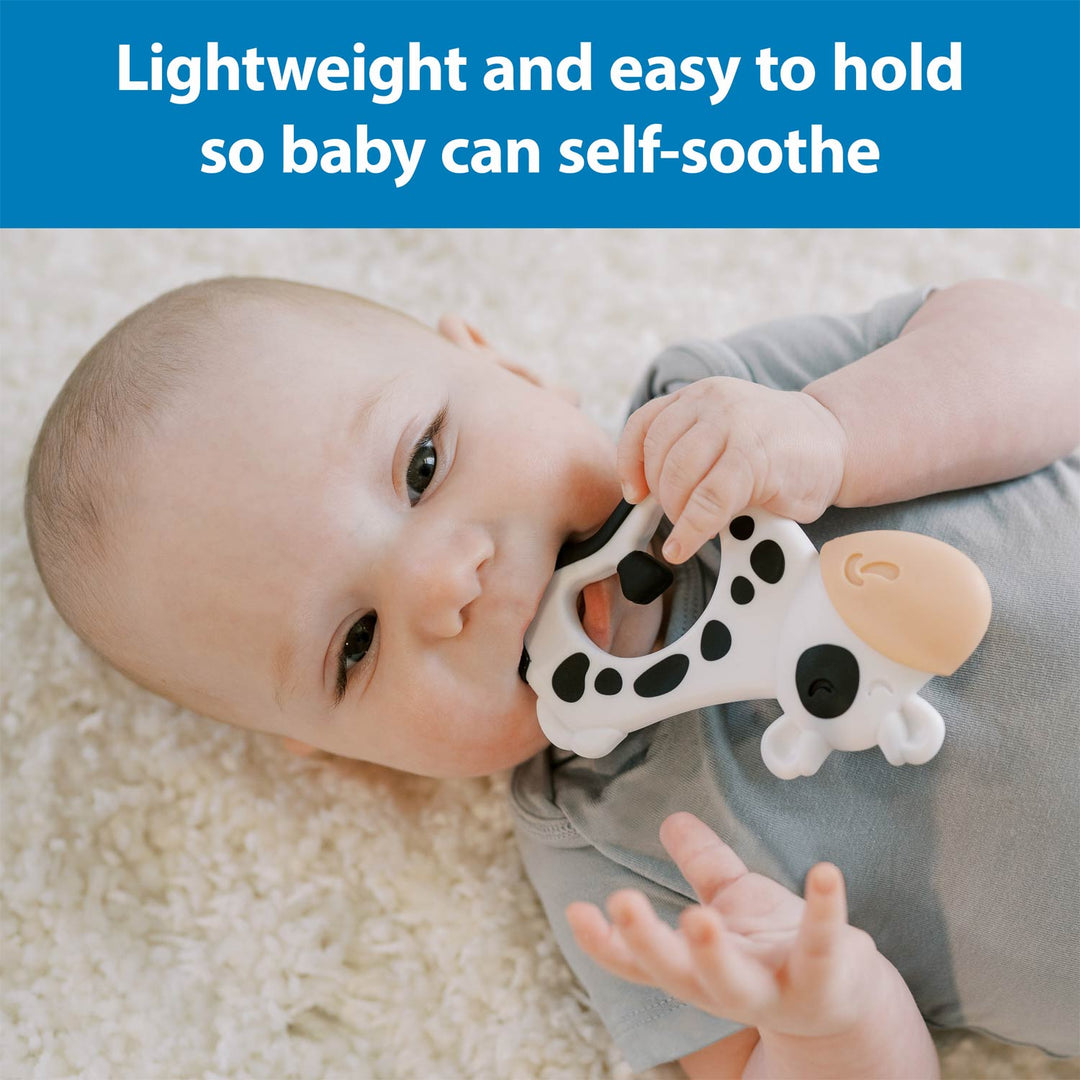 A baby lying on a cream textured blanket holds a cow-shaped teether toy with a blue text banner at the top that reads 'Lightweight and easy to hold so baby can self-soothe'.