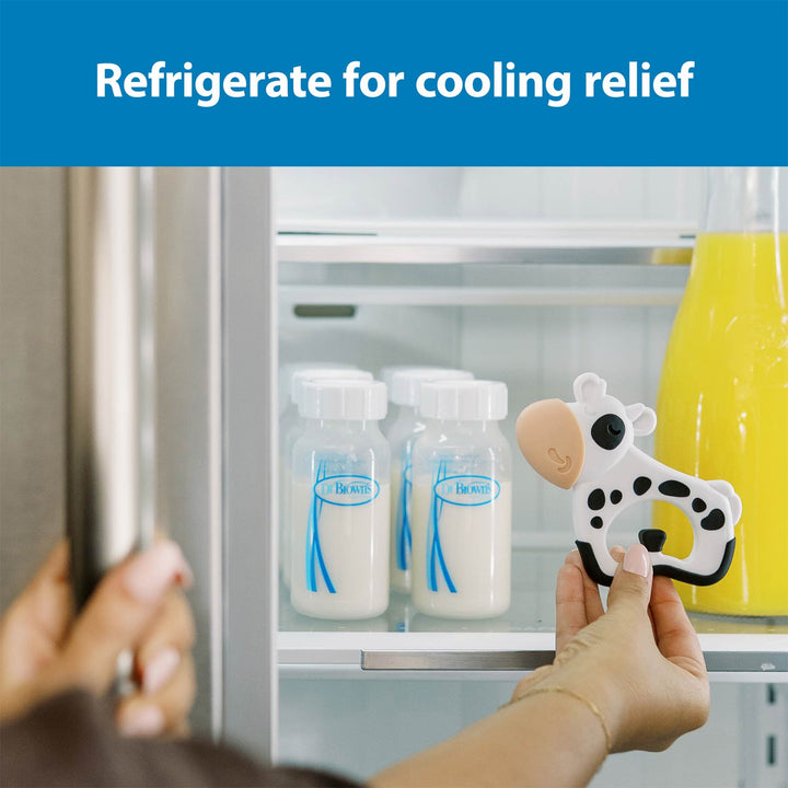 A person holds a cow-shaped refrigerator teether inside an open refrigerator shelf next to milk bottles and a yellow bottle, with the text 'Refrigerate for cooling relief' at the top.