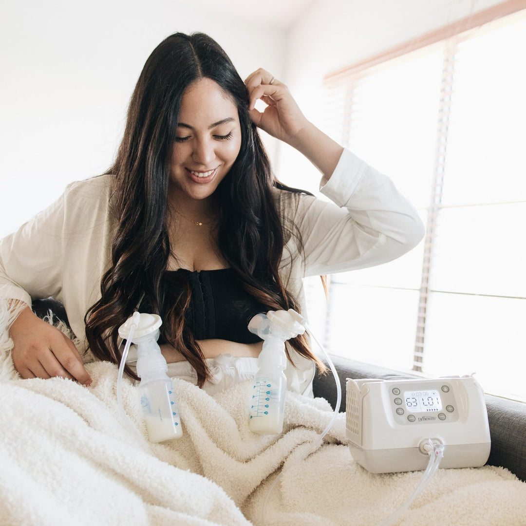 A woman with long dark hair sits comfortably with a light blanket, using a breast pump. Two bottles are attached to the pump, which rests on her lap. She wears a white shirt and smiles softly in a bright room with window blinds.