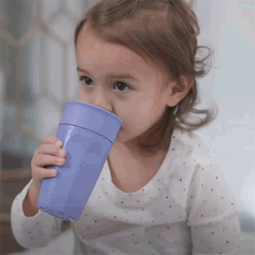 A young child with light brown hair drinks from a large, blue Dr. Brown’s® Milestones™ Cheers360™ Cup (10 oz/300 mL) using both hands. The child, in a white shirt with small colorful dots, looks to the side.