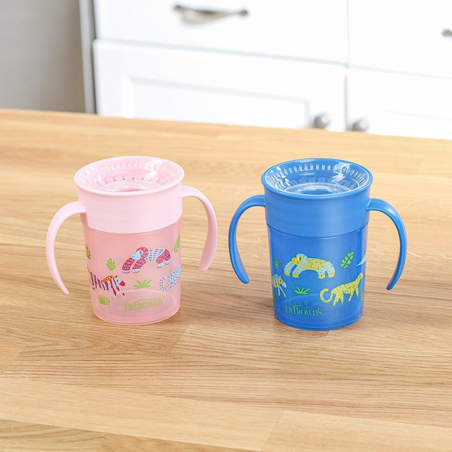 Two colorful children's sippy cups with handles on a wooden countertop. One is pink with playful animal designs, and the other is blue with similar patterns. White kitchen cabinets are visible in the background.