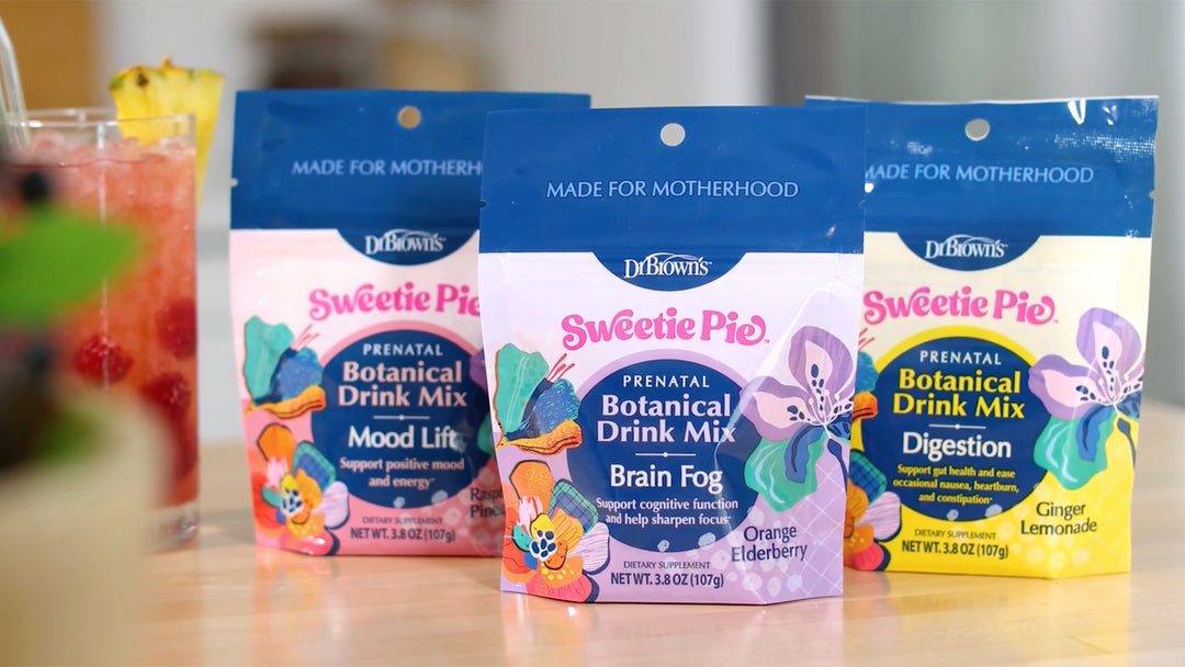Three Dr. Brown's Sweetie Pie™ Prenatal Botanical Drink Mix bags lined up on a wooden surface, with a glass of pink drink in the blurred background.