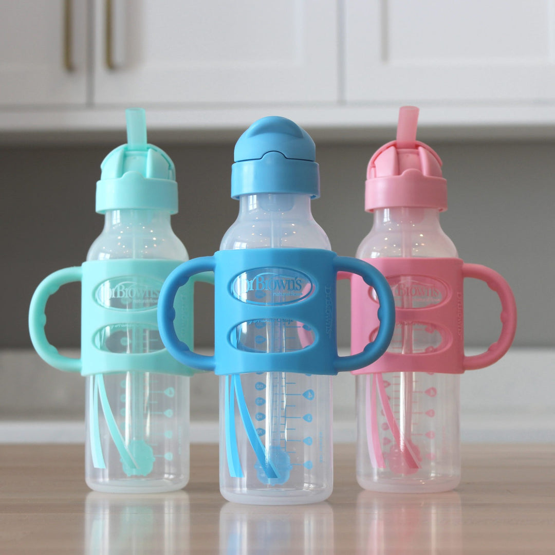 Three baby bottles with handles in front of a neutral kitchen background. Each bottle has a built-in straw and colorful lids: one mint green, one blue, and one pink.