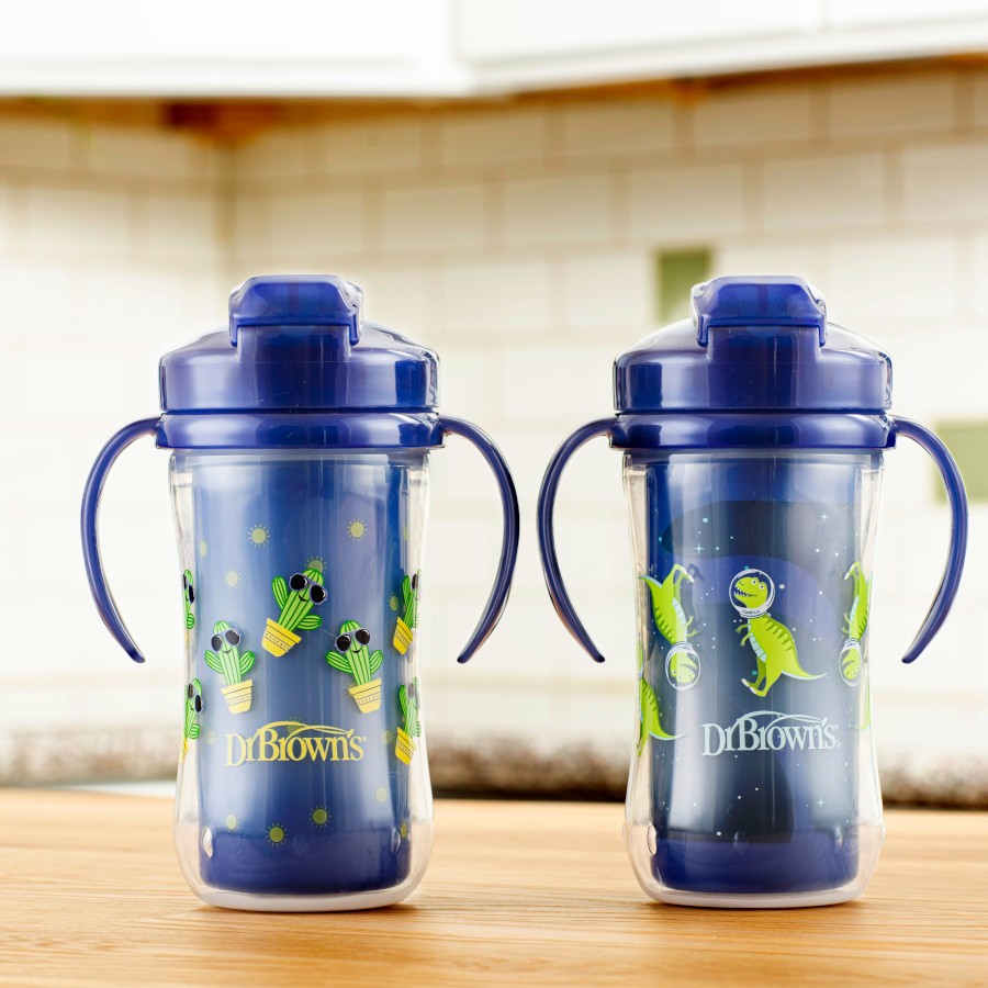 Two Dr. Brown's toddler cups with handles are on a kitchen counter. They feature playful dinosaur and cactus designs on a clear background. The lids and handles are a matching shade of blue. The cups stand against a neutral kitchen tile backdrop.