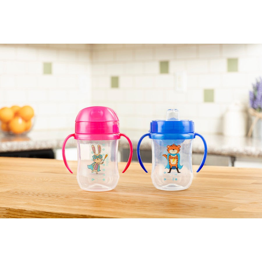 Two baby sippy cups on a wooden counter. The pink cup has a bunny design and dual handles, while the blue cup features a tiger, also with dual handles. A fruit bowl and lavender vase are blurred in the background.