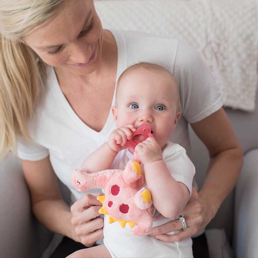 A woman holding a baby who is chewing on a pink pacifier attached to a dinosaur plush toy. The woman and baby are sitting on a chair, and there is a white blanket in the background. Both are wearing white clothing.