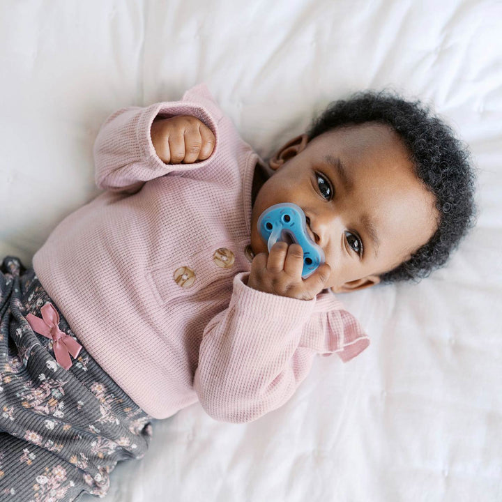 A baby with soft skin wears a pink long-sleeve shirt and patterned pants, lying on a white blanket while holding a Dr. Brown’s® Advantage™ One-Piece Silicone Pacifier from Dr. Brown’s near their mouth and looking up.