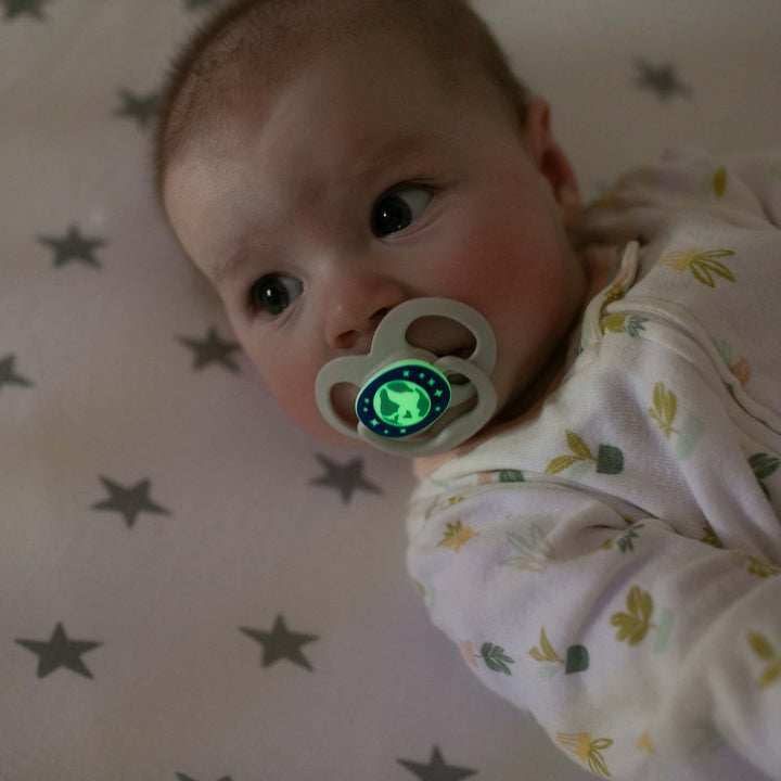 A baby rests on a white starry sheet, wearing a pale onesie with leaf patterns, looking to the side while comforted by Dr. Brown's™ Advantage™ Glow-in-the-Dark Pacifier.