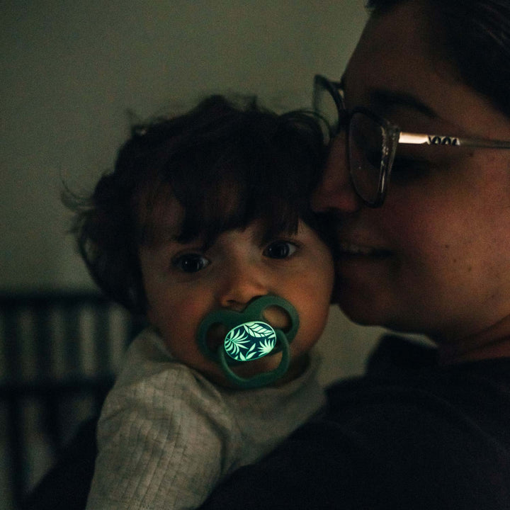 A young child with Dr. Brown's™ Advantage™ Glow-in-the-Dark Pacifier is held by a glasses-wearing adult in a dimly lit room, creating a cozy atmosphere. The child's wide eyes and dark, curly hair enhance the tender moment.