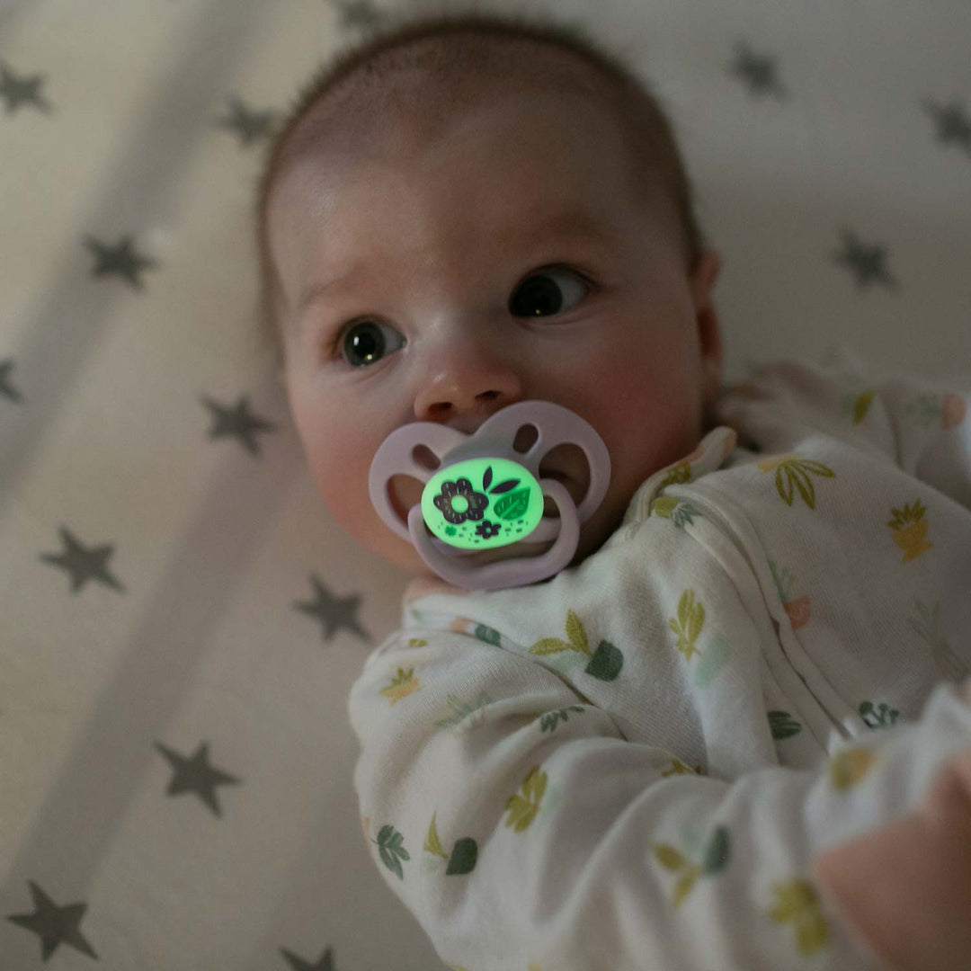 A baby rests on a star-patterned sheet, with Dr. Brown's™ Advantage™ Glow-in-the-Dark Pacifier in their mouth. This symmetrical soother emits a gentle green light with floral designs. The baby wears a light-colored outfit sprinkled with small prints.