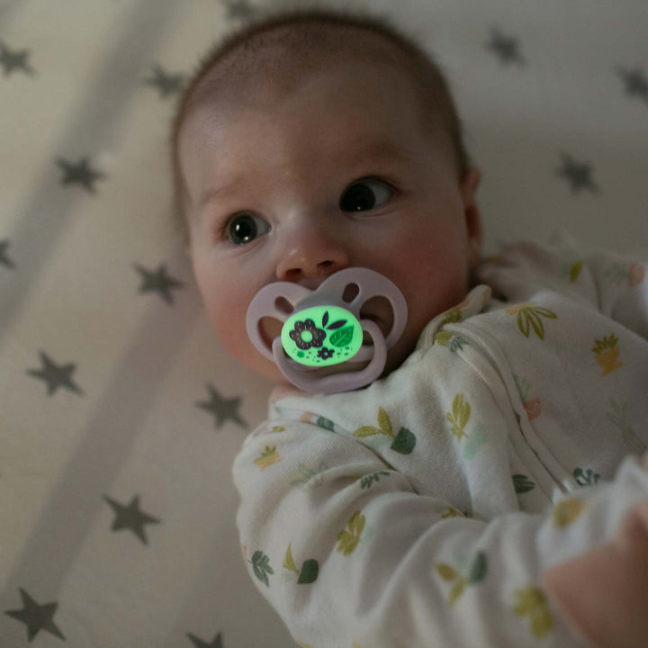 A baby rests on a star-patterned sheet, with Dr. Brown's™ Advantage™ Glow-in-the-Dark Pacifier in their mouth. This symmetrical soother emits a gentle green light with floral designs. The baby wears a light-colored outfit sprinkled with small prints.