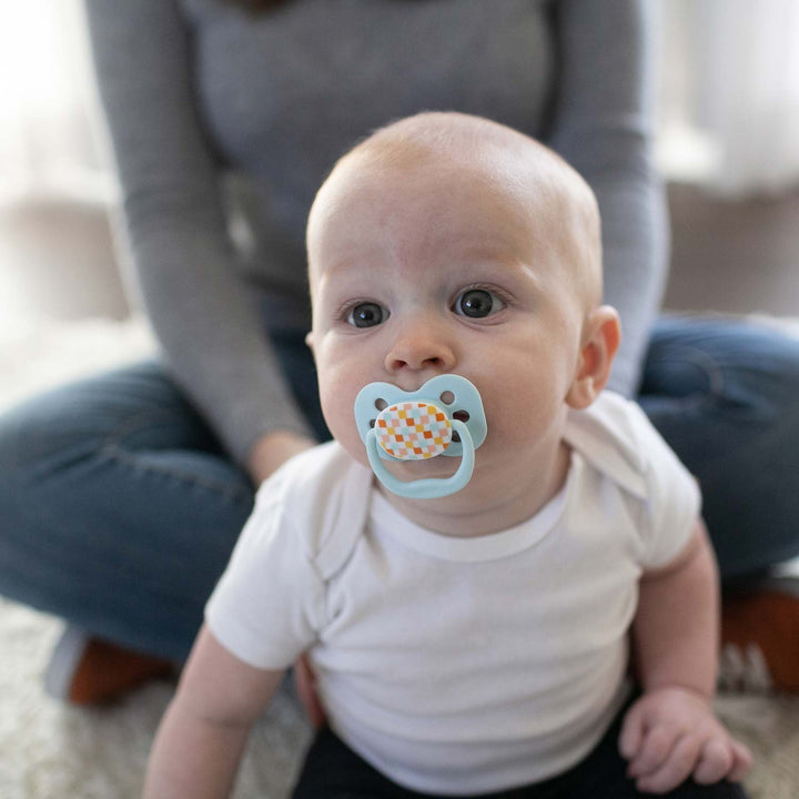 A baby sitting in front of a parent, with a light green Advantage pacifier with a checkered pattern on it