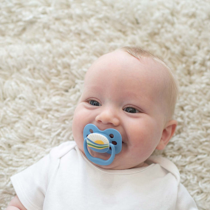 A baby lying on the ground with a blue Advantage pacifier with a sunrise over water on it