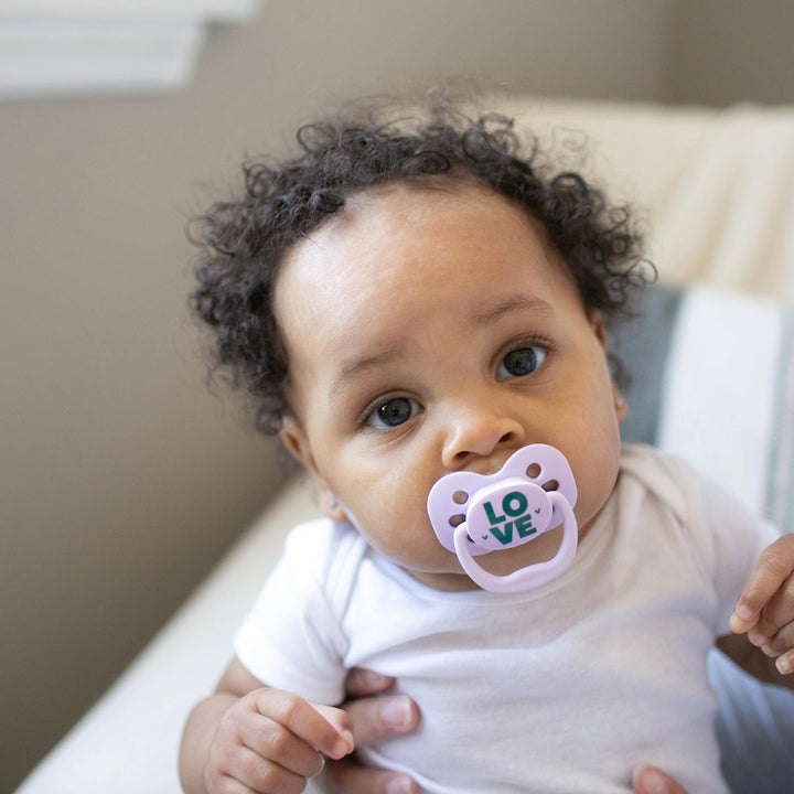 Curly-haired baby with a pink Advantage pacifier that says LOVE