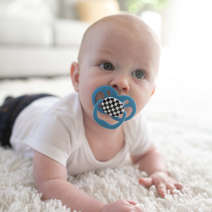 A baby in a white onesie curiously holds its head up while using a Dr. Brown’s™ Advantage™ Pacifier, featuring a black and white checkered pattern, on a soft carpet. The softly blurred background suggests a bright, cozy room.