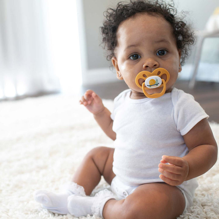 A curly-haired baby sits on a soft white carpet, wearing a white onesie and socks, enjoying a bear-shaped Dr. Brown's™ Advantage™ Pacifier from the 2-count pack, featuring a reversible bulb with a yellow circular center.