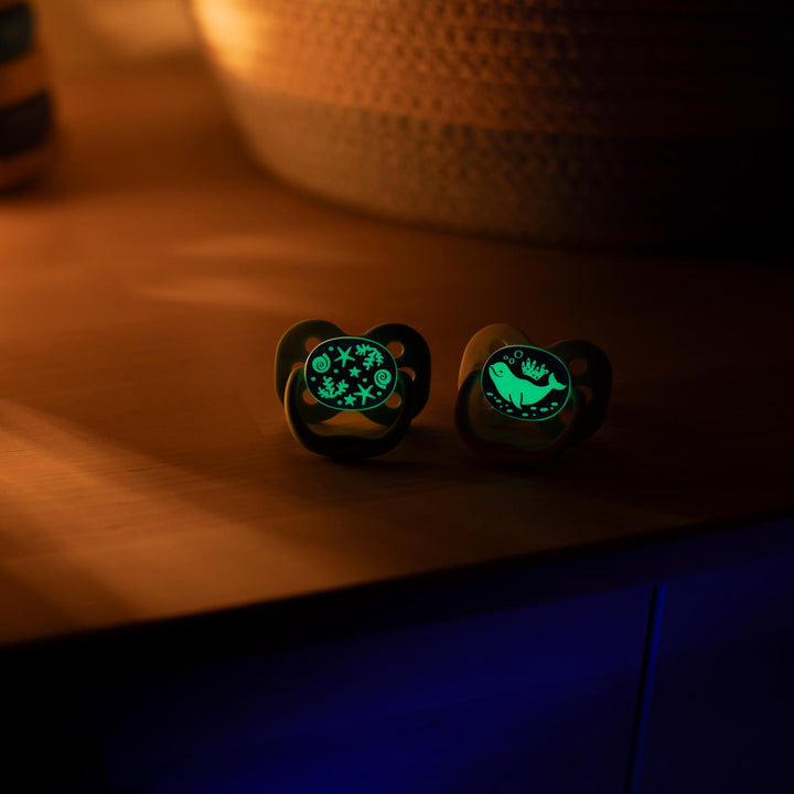On a wooden surface, two Dr. Brown's™ Advantage™ Glow-in-the-Dark Pacifiers sit glowing warmly—one adorned with stars and planets, the other featuring a crowned dinosaur—enhancing their charm and creating a cozy atmosphere.