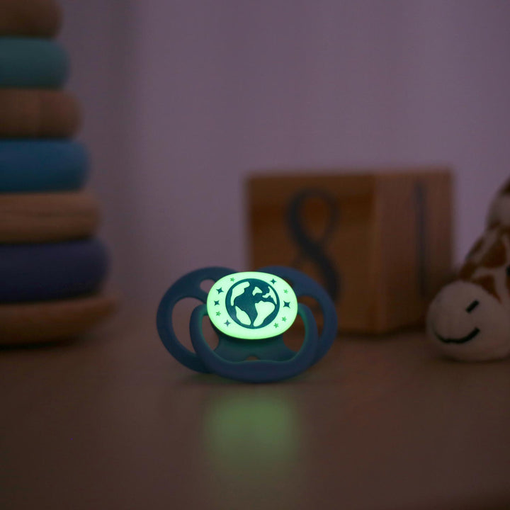 A Dr. Brown's™ Advantage™ Glow-in-the-Dark Pacifier with a planet design rests on wood, glowing softly. Blurred in the background are a wooden stacking toy, a plush toy, and a block with the number 8.