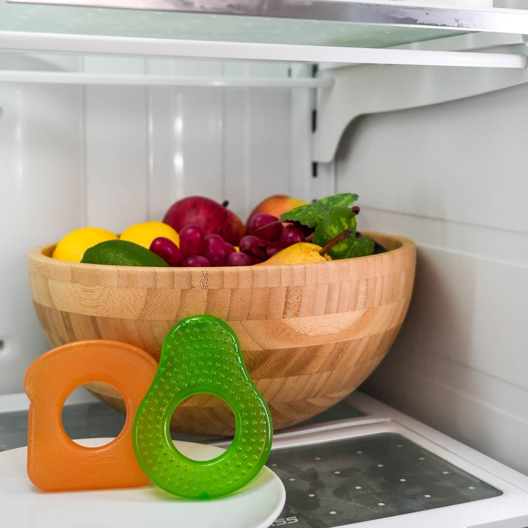 A wooden bowl of assorted fruits is in the fridge. In front are two Dr. Brown's® AquaCool Water-Filled Teethers – Avocado Toast, perfect for soothing a teething baby.