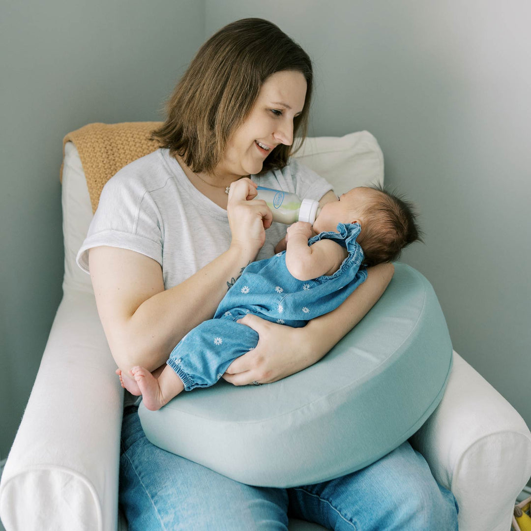 Woman feeding a baby with a bottle while sitting with a green Feeding and Nursing Lap Pillow on her lap