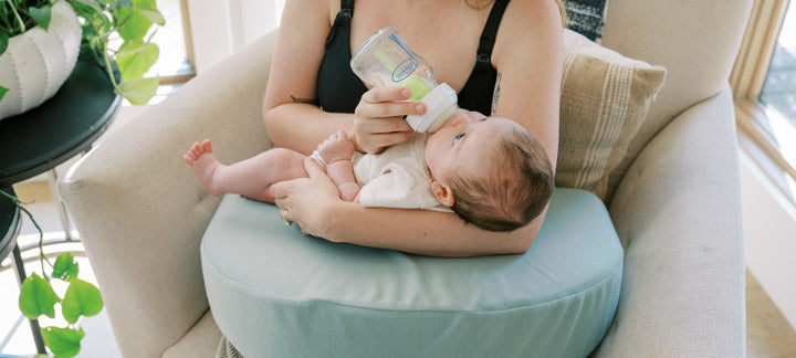 Woman holding a baby and bottle feeding it on a cushioned chair with a Feeding and Nursing Lap Support Pillow