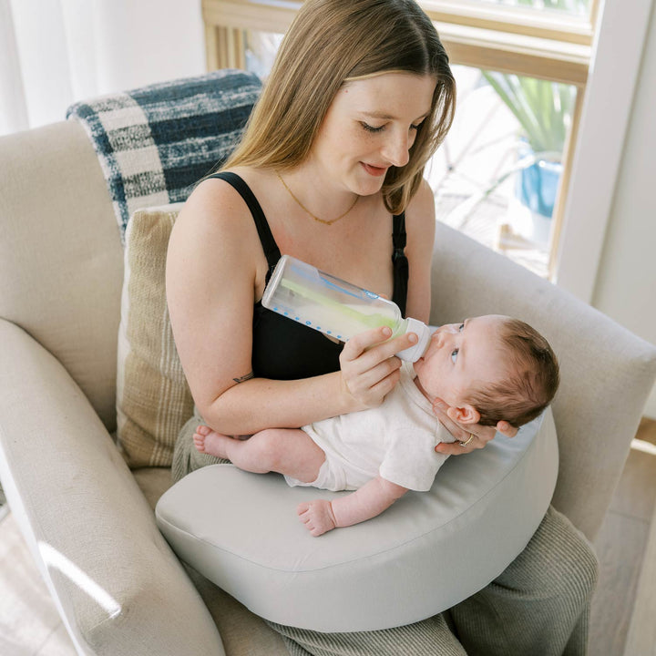 Woman feeding a baby with a bottle while sitting with a gray Feeding and Nursing Lap Support Pillow on her lap
