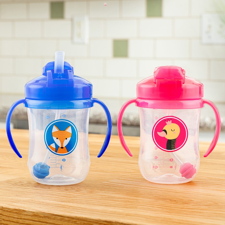 Two sippy cups with animal designs on a wooden counter. The blue cup has a fox design, and the pink cup features a duck. Both have handles and spouts. A tiled kitchen backsplash is visible in the background.