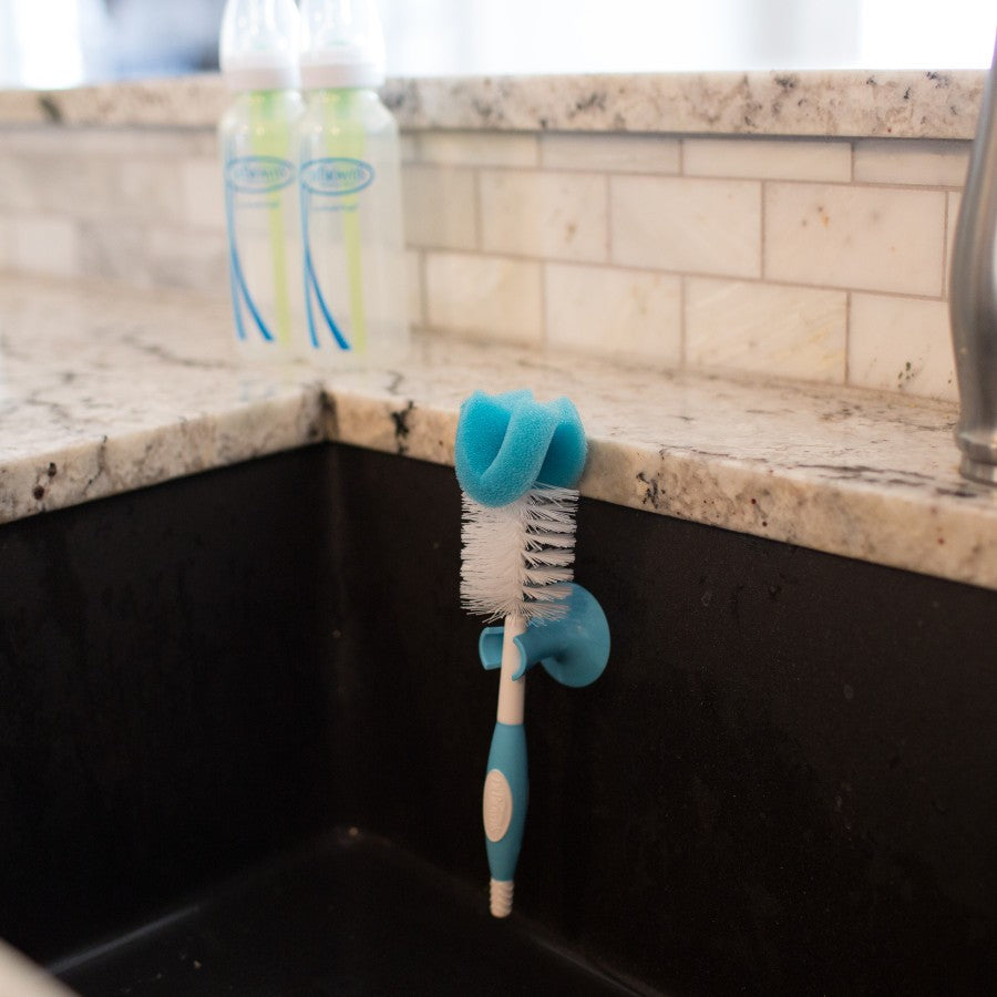 A blue and white bottle cleaning brush with a suction cup attached to the edge of a kitchen sink. Two baby bottles are in the background, placed on a marble countertop. The backsplash features white subway tiles.