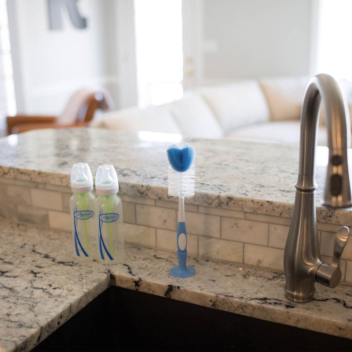 Two baby bottles and a Dr. Brown's™ Baby Bottle Brush with a bristle and sponge head are placed on a kitchen countertop next to a sink with a modern faucet.