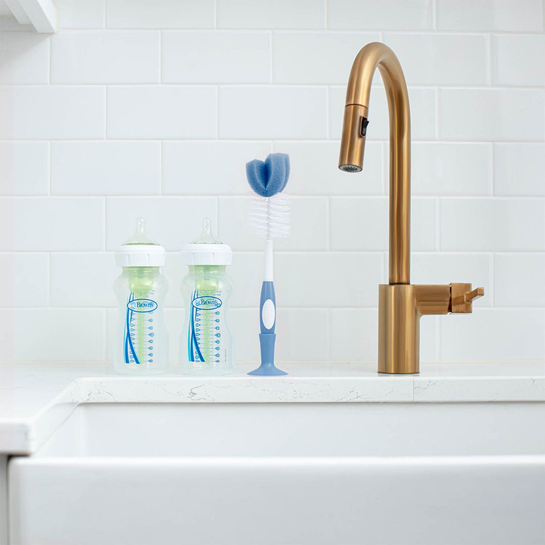 Gold faucet on a white kitchen sink with baby bottles and a brush against a tiled wall.