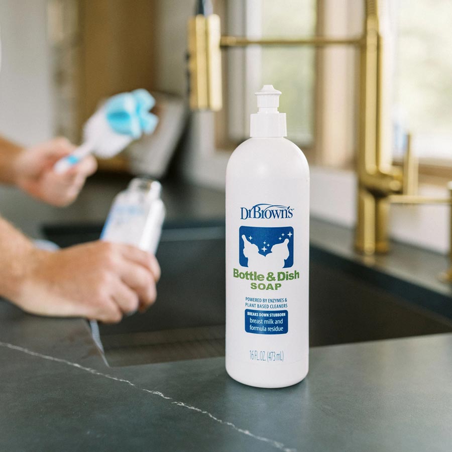 A bottle of Dr. Brown's Bottle & Dish Soap is placed on a kitchen counter. In the background, a person is washing a bottle at the sink. The soap bottle is white with blue and green labeling.