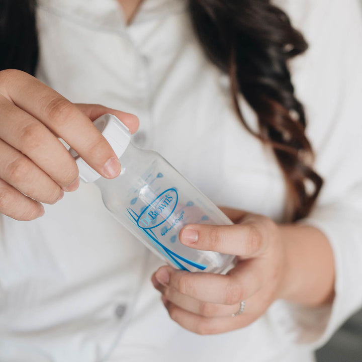 A person in a white shirt opens a clear Dr. Brown's bottle with a white Dr. Brown’s Natural Flow® Storage/Travel Cap Narrow, 3-Pack; only their hands and torso are visible.