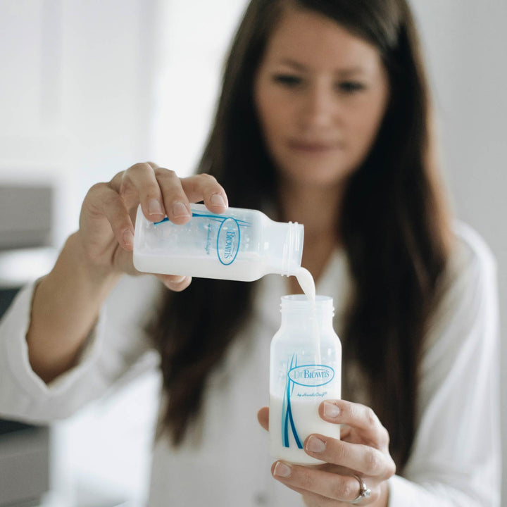 A woman in a white shirt pours milk from one Dr. Brown's™ Breast Milk Collection Bottle into another, with a blurred background.