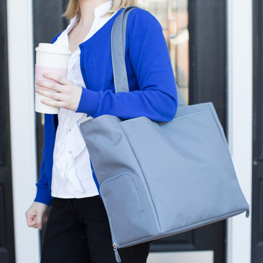 Person in a blue cardigan holding a large gray bag and a takeout coffee cup, standing in front of a dark door.